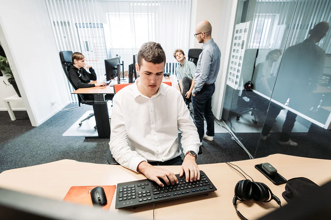 An Infodatek Group engineer at his desk, with colleagues talking in the background.