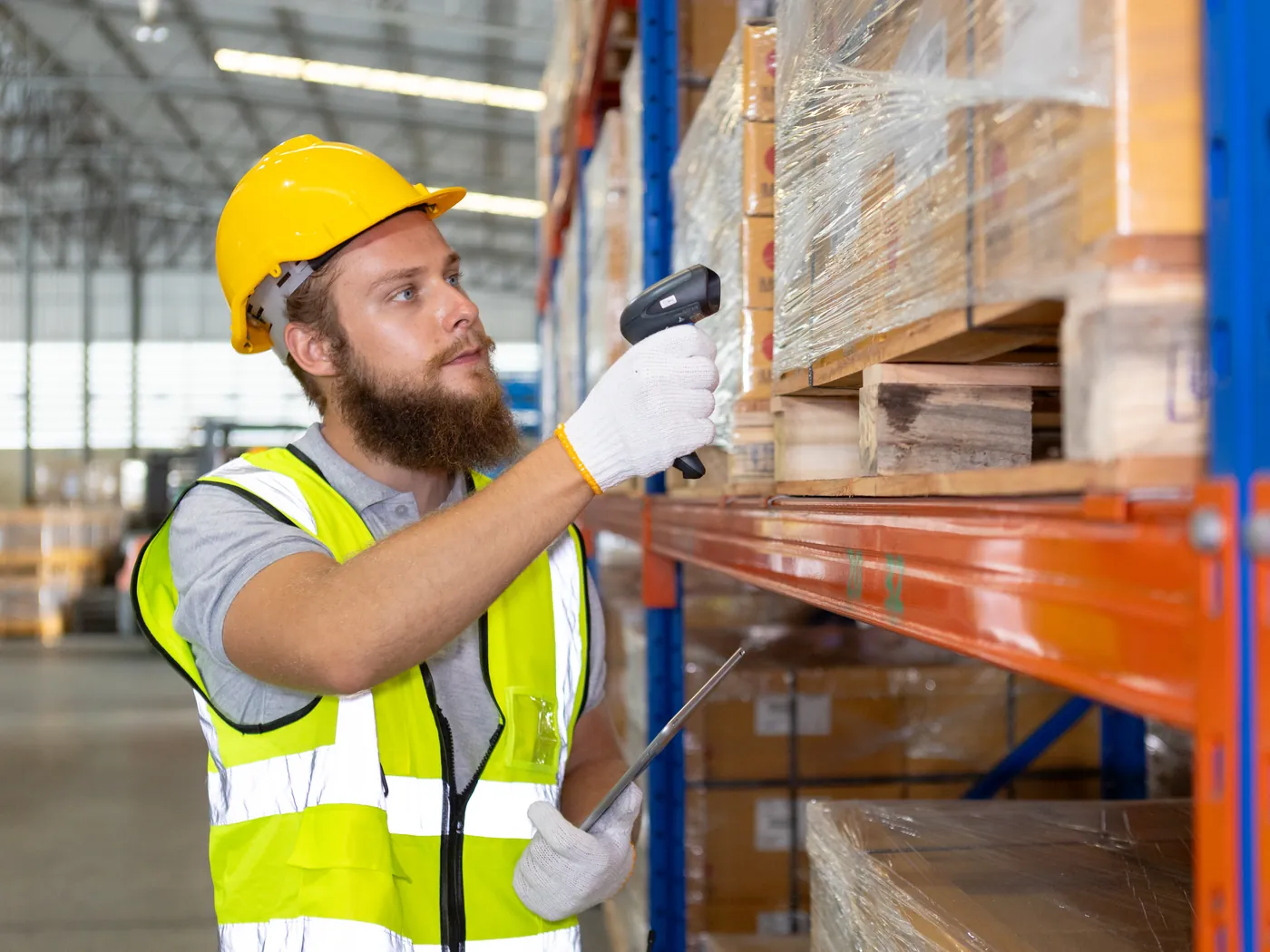 A warehouse worker in a hard hat and vest scans a box on the racking.