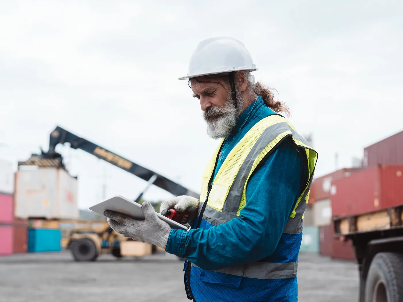 A worker in a hard hat checks a tablet at a container terminal, with a reach stacker in the background.