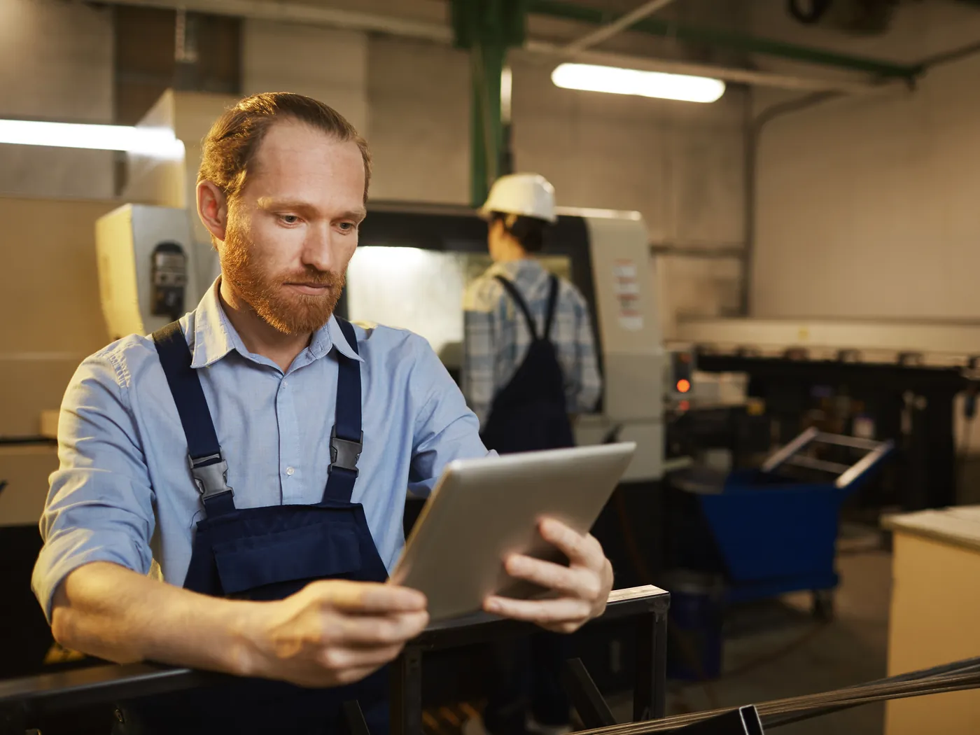A technician in work overalls looks at a tablet in a production hall with machines.