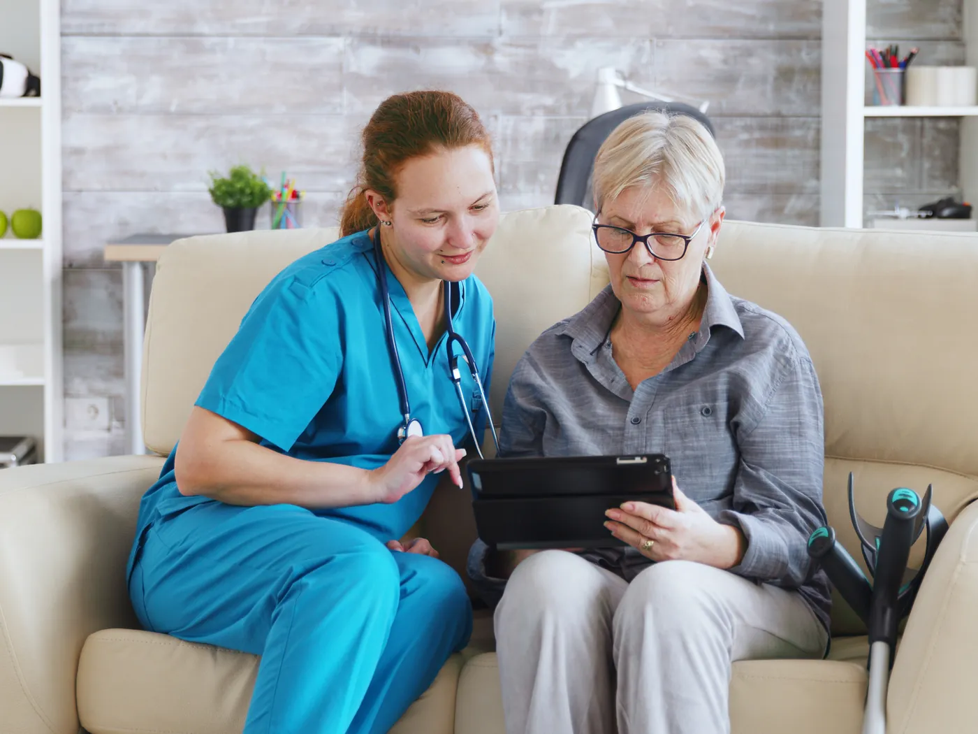 A care worker shows an older woman something on a tablet on the sofa.